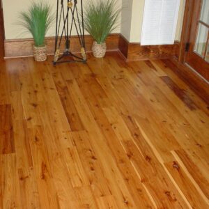 Polished honey-toned hardwood floor stretching across a room with two wicker potted ornamental grasses beside a black metal plant stand, a white wall vent, and a wooden glass‑pane door at the right.