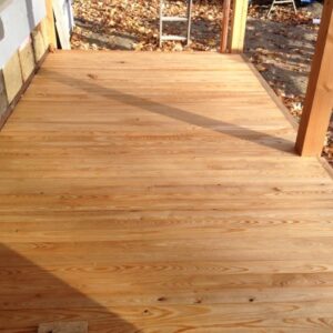 Newly installed light-wood deck with parallel planks and visible grain, vertical support posts casting long shadows and fallen leaves and a ladder visible beyond the deck near the house wall.