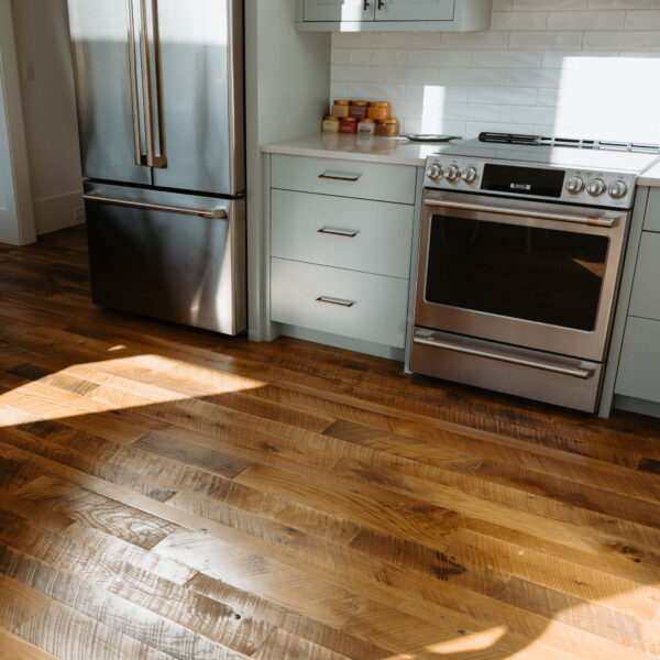 Sunlit modern kitchen corner with a stainless-steel French-door refrigerator and matching range beneath a microwave, light-gray cabinets and drawers, small jars on the countertop, and warm hardwood floors with sunlit patches and shadows.