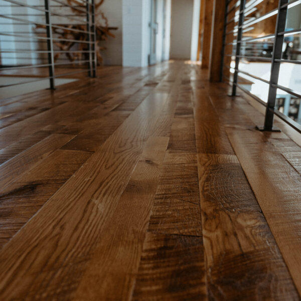 Low-angle close-up of a warm-toned hardwood floor stretching down a hallway with horizontal metal railings on both sides, exposed wooden beams and a doorway visible in the distance.