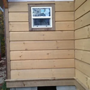 Square log siding Exterior wall of a small wooden building with horizontal light-brown plank siding, a small white-framed rectangular window near the top with dark brown trim, corner boards at the edge, and a raised concrete-block foundation with an open crawlspace below.