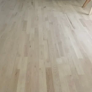 Maple Flooring Light natural hardwood plank floor extending toward a back wall with a white cushioned bench and table legs visible near an open doorway.