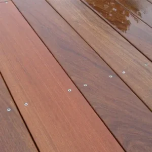 Ipe Wood Decking Close-up of reddish-brown wooden deck planks showing wood grain and a row of evenly spaced metal screw heads, with a small wet reflective patch at the top edge.