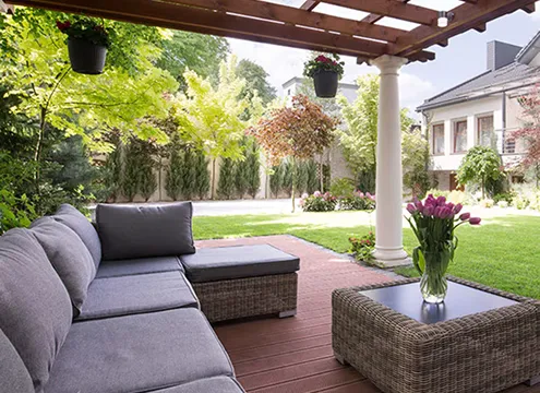 Covered wooden pergola patio with a gray wicker sectional sofa and matching ottoman, a low wicker coffee table topped with a vase of purple tulips, overlooking a manicured lawn, trees, and a house in the background.