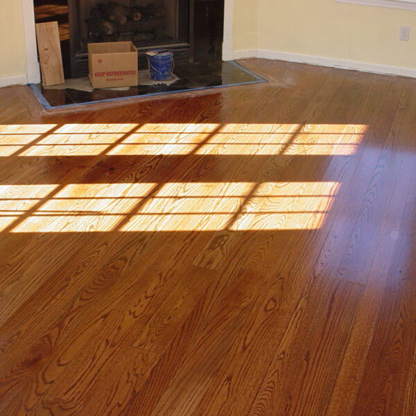 Polished hardwood floor lit by rectangular window-pane shadows, with a fireplace hearth in the background containing a cardboard box and a paint bucket.