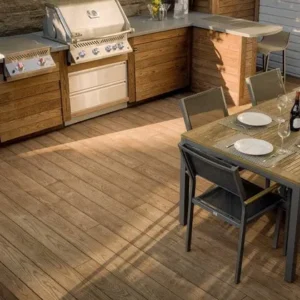 Thermally Modified Decking Sunlit outdoor wooden-deck kitchen and dining area featuring a built-in stainless-steel grill in wood cabinetry with a stone countertop and bar stools, and a nearby table set with plates, a wine bottle and chairs.
