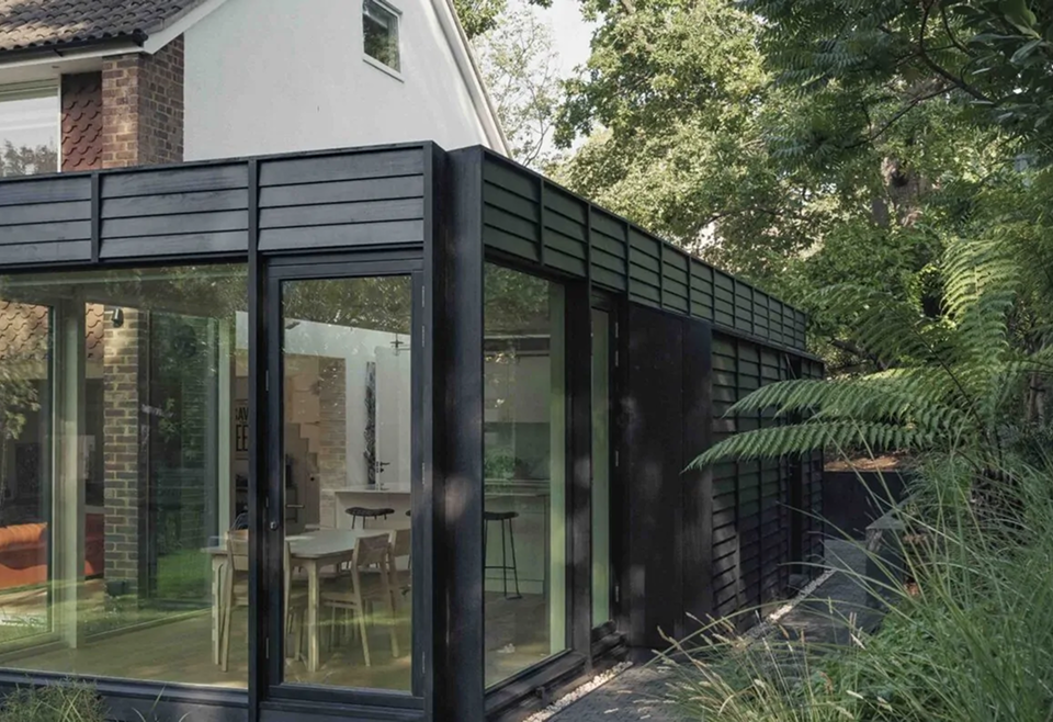 Modern black-framed glass extension attached to a brick house, showing a dining table and chairs through floor-to-ceiling windows, with dark horizontal cladding and dense trees and ferns surrounding the structure.