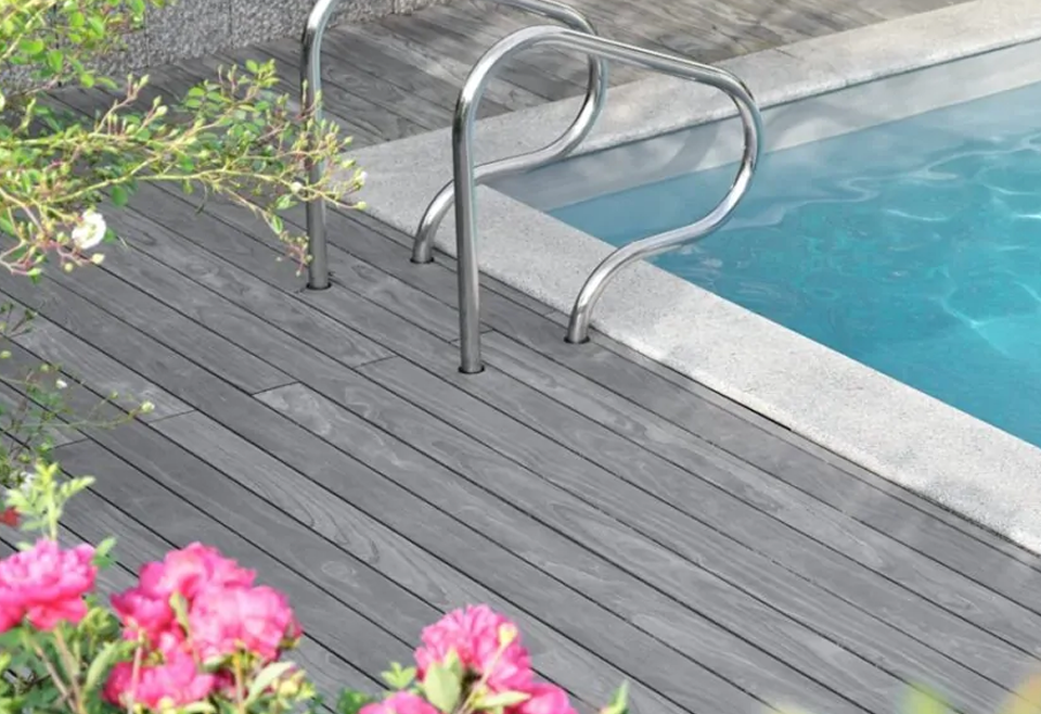 Corner of a swimming pool with clear blue water, a stainless-steel ladder set in light concrete coping, a gray wooden deck, and pink flowering shrubs in the foreground.