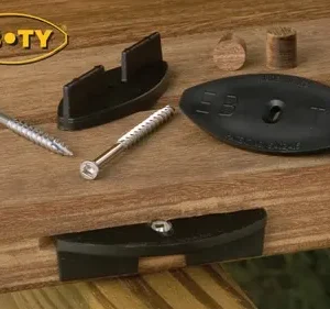 EB-TY branded concealed deck fastener kit laid out on a wooden deck board, showing a black oval spacer plate, a curved black clip, two screws and two wooden plugs with the EB•TY logo in the corner.