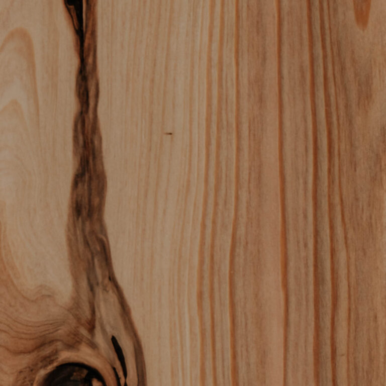 Close-up of a light-brown wooden plank with vertical grain lines and a dark knot and streak near the lower-left corner.