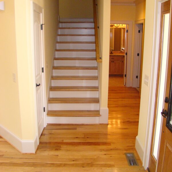 Hallway entry with a central straight staircase featuring white risers and wooden treads, honey-toned hardwood floors, pale yellow walls, and a doorway leading to a bathroom down the hall.