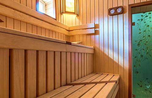 Sunlit wooden sauna interior with tiered slatted benches and vertical wood paneling, a wall-mounted thermometer/hygrometer, and a green frosted glass door patterned with leaves.