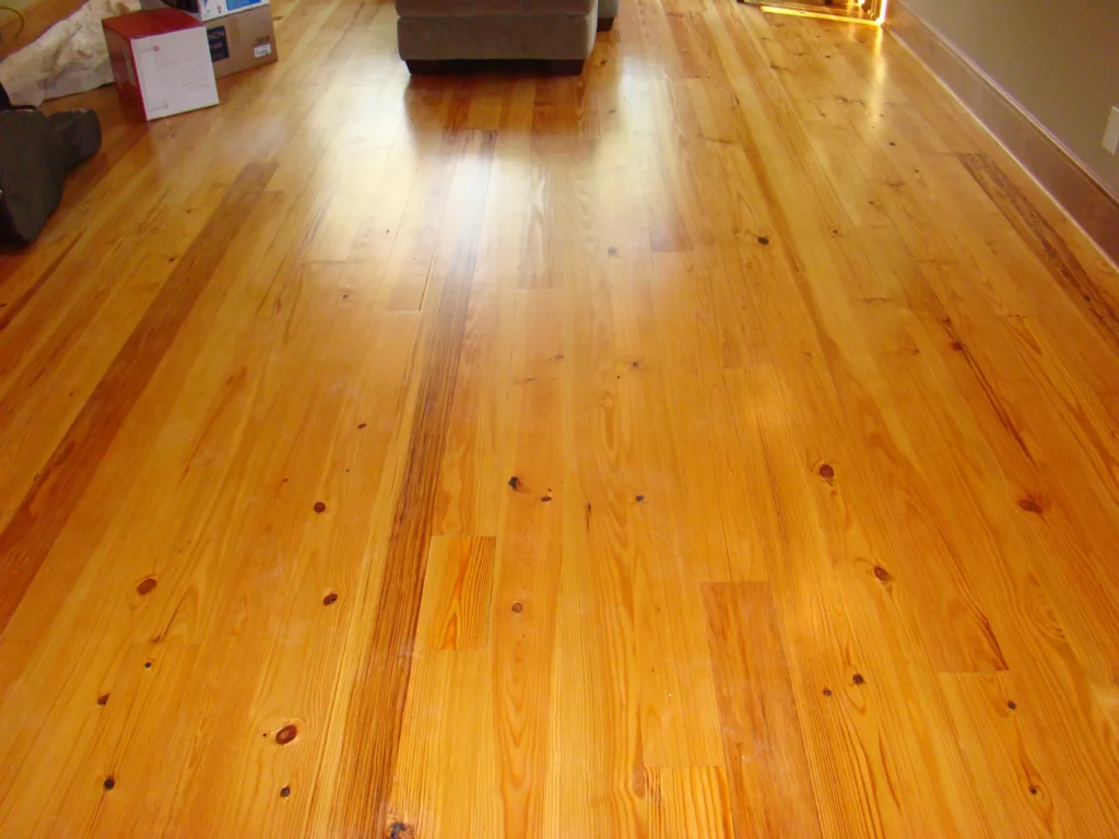Honey-colored hardwood floor with visible knots and grain running toward a gray sofa at the far end and a few cardboard boxes near the left wall.