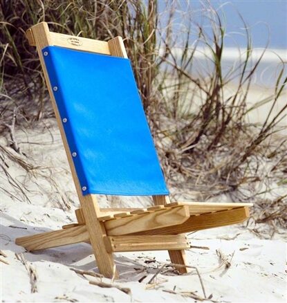 Low wooden beach chair with bright blue fabric set on white sand near dune grasses, with four small color swatches (navy, red, dark green, slate blue) along the bottom.