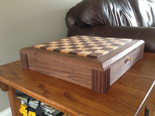 Raised wooden chessboard box with alternating light and dark inlaid squares, brass drawer knob and decorative corner accents, sitting on a polished wooden side table with a brown leather sofa behind it.