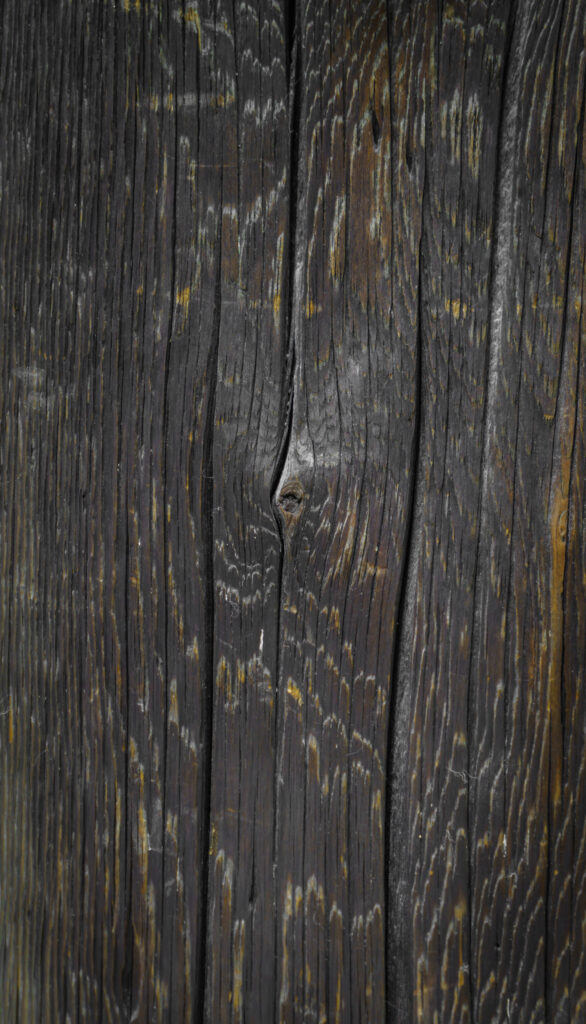 Close-up of vertically arranged weathered dark wooden planks showing deep grain lines, a central knot and cracks, with streaks of lighter gray and yellowish discoloration.