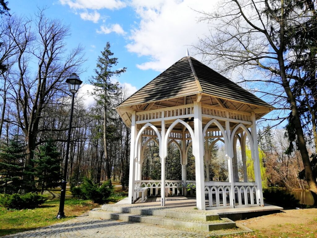 White wooden gazebo with a pointed shingled roof and Gothic-style arched supports on a low stone platform by a cobblestone path, framed by leafless trees and a black lamppost under a blue sky.