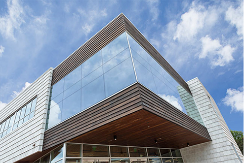 Low-angle view of a modern corner building with floor-to-ceiling glass panels reflecting a blue sky, a wood-paneled overhang, and light-colored stone cladding.