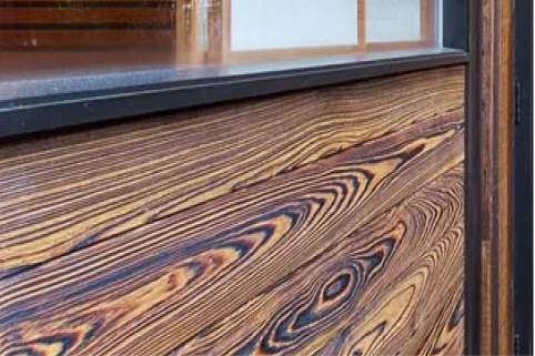 Close-up of a wooden wall panel showing bold, swirled dark and light grain patterns beneath a black window sill and dark metal frame.