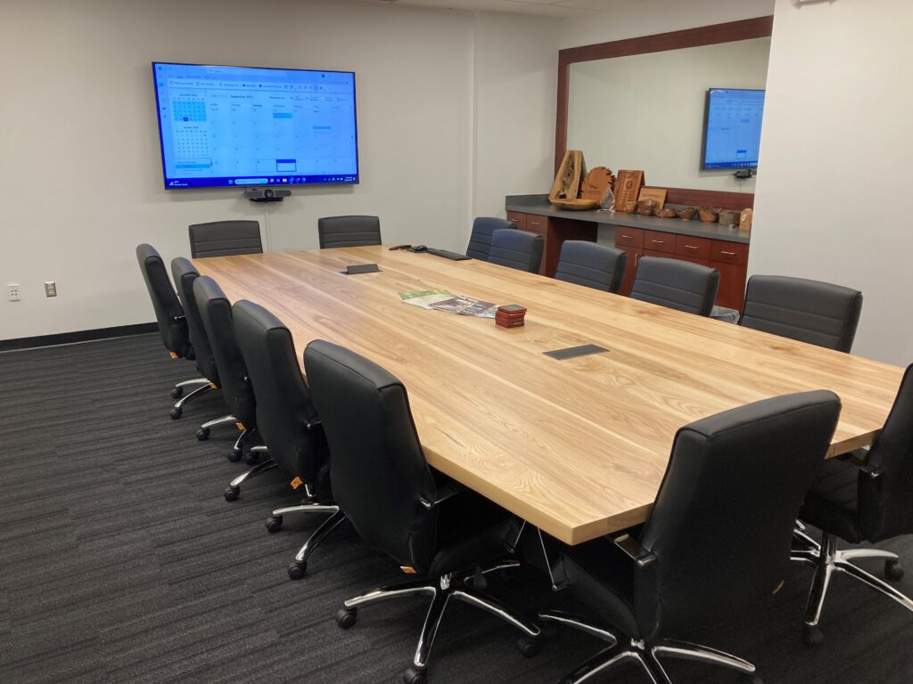 Long light-wood conference table surrounded by black leather swivel chairs in a modern meeting room with wall TV.