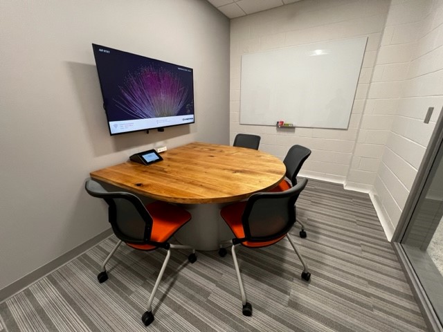 Small meeting room: round wood table, four gray chairs with orange seats, wall-mounted monitor and whiteboard.