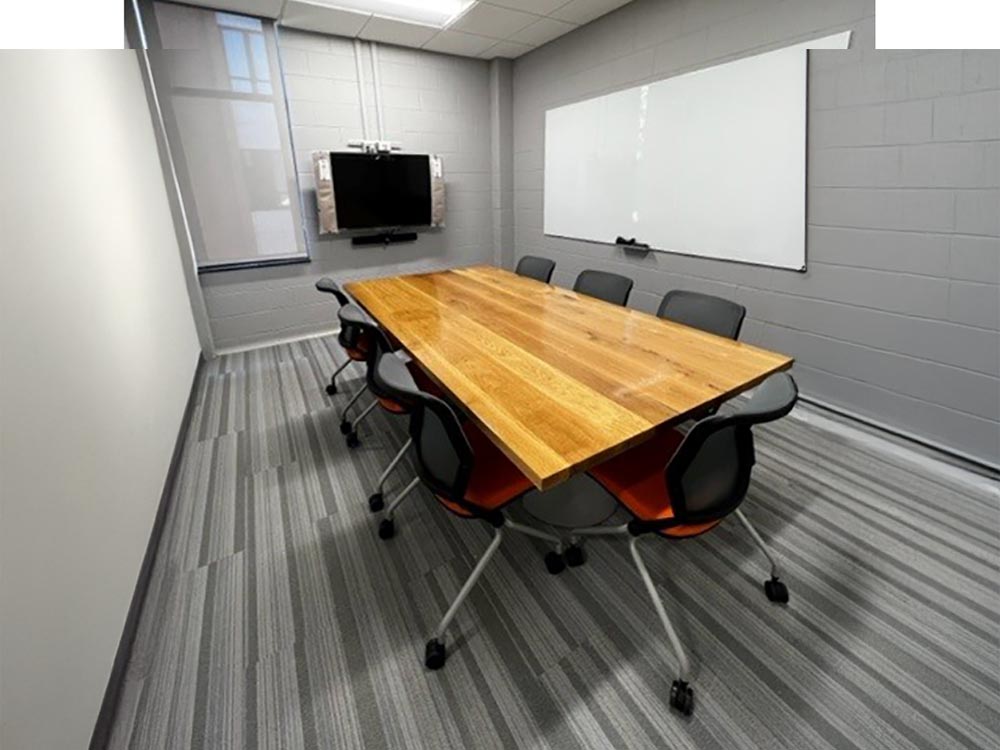 Small conference room with long wooden table surrounded by black-and-orange rolling chairs, whiteboard and wall-mounted TV.