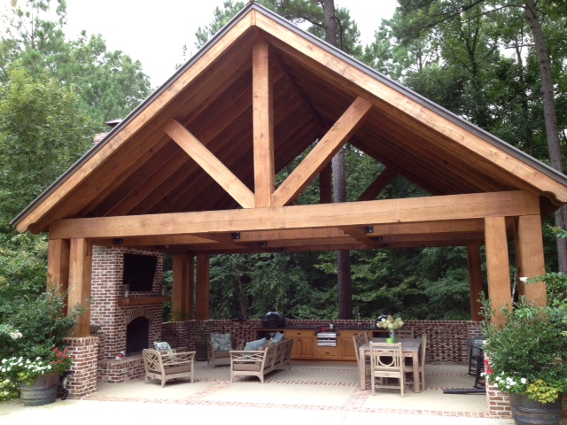 Timber-framed open pavilion with peaked gabled roof, exposed beams, brick fireplace, and patio seating and dining amid trees