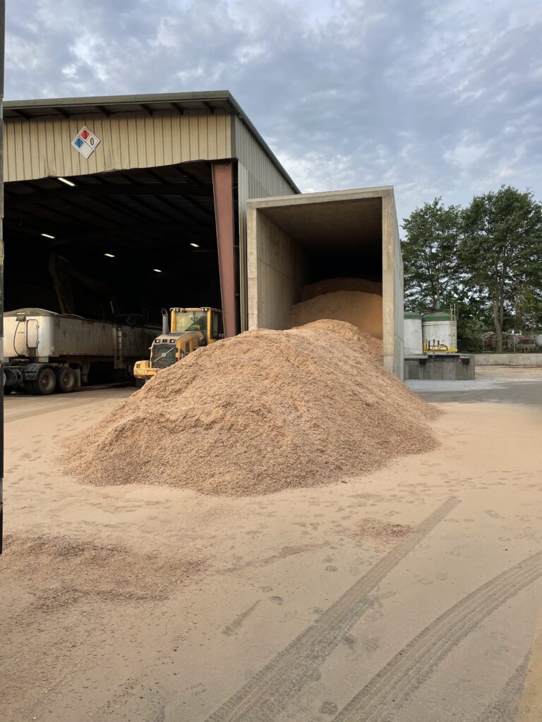 Large pile of light-brown sawdust in front of an open industrial storage bay with trucks and trees in background